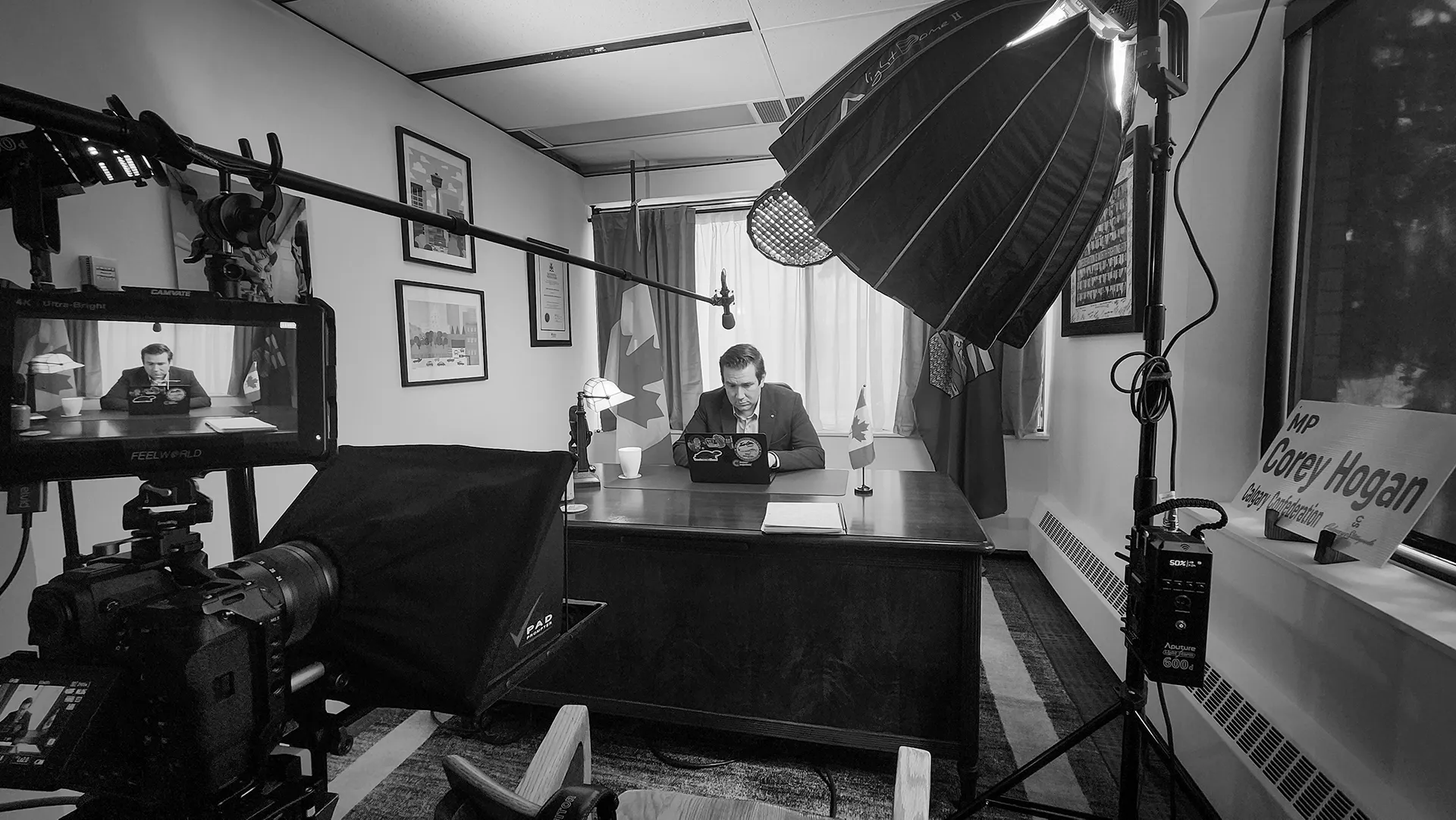 Black and white photo of MP Corey Hogan rewriting his script at his desk in his constituency office, surrounded by video production equipment including a teleprompter, boom microphone, and softbox lighting, during the rapid-response shoot for his Alberta separatism video.